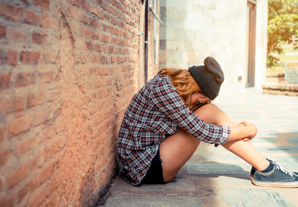 A light-skinned femme person with blond hair wearing a plaid shirt, black shorts, sneakers, and a beanie sits against a brick wall hugging their knees with their head down.