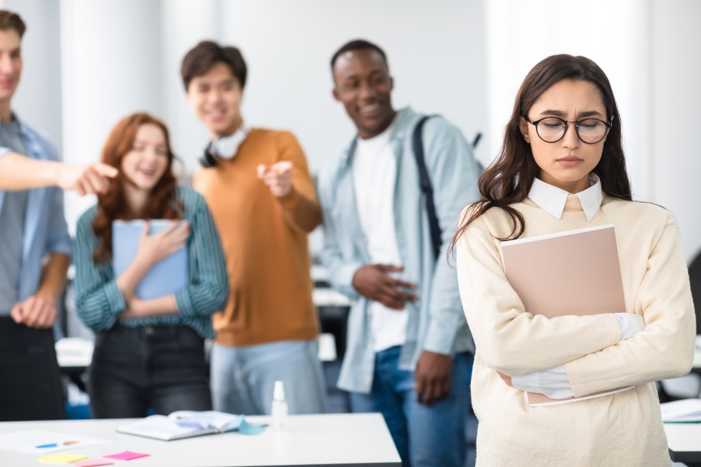 A sad Latin woman in eyeglasses standing away alone, suffering humiliation and public disgrace, a crowd of people bullying pointing fingers at the victim, laughing. 