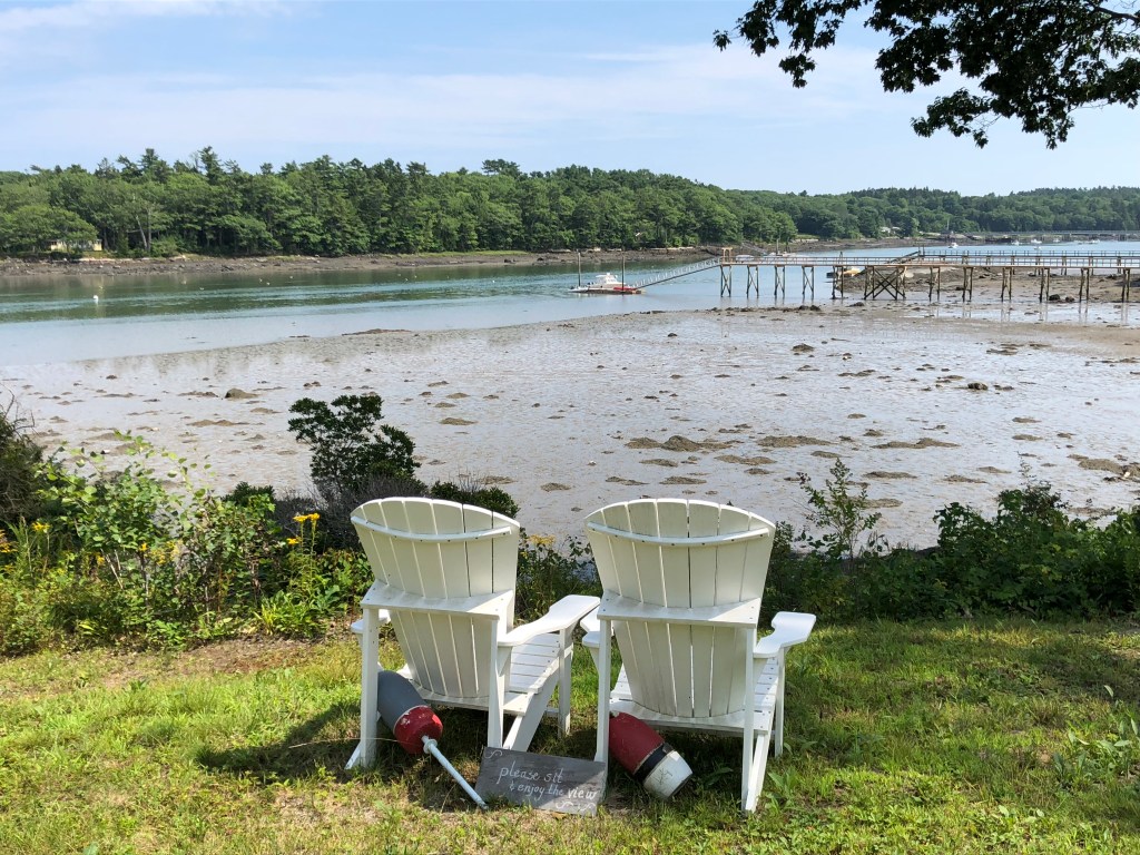 Photo taken from a river bank in Maine. Two white Adirondack chairs are in the foreground with the river at low tide and a residential dock in the background.
